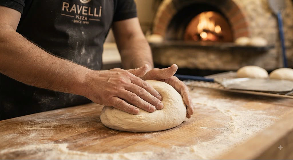 "Ravelli Pizza — since 1999" yazılı önlüğüyle bir usta, taş fırının önünde elle hamur yoğuruyor.